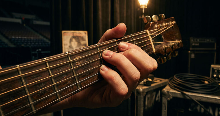 Calloused worker hand on acoustic strings backstage, representing Nancy Wilson’s hidden arena guitar labor